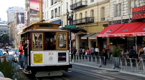 A vintage cable car travels down a city street, with shops and colorful decorations lining the sidewalk.