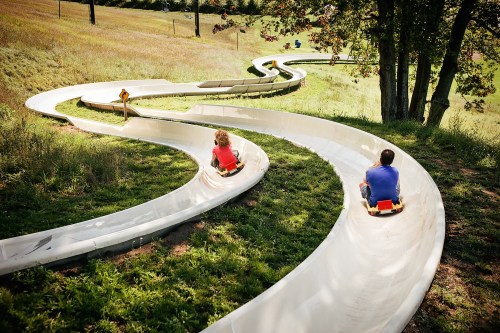 Two children ride down a winding slide through a grassy landscape on a sunny day.