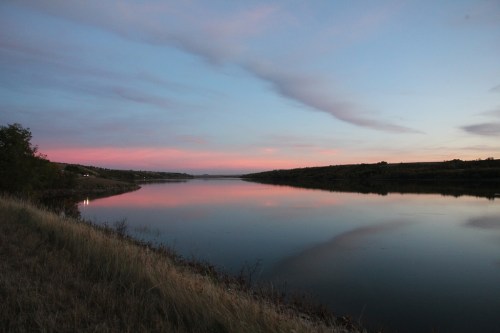A serene river at dusk, reflecting soft pink and blue hues in the sky, surrounded by grassy banks.