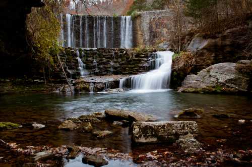 A serene waterfall cascades over rocks into a calm pool, surrounded by autumn foliage and a stone wall.