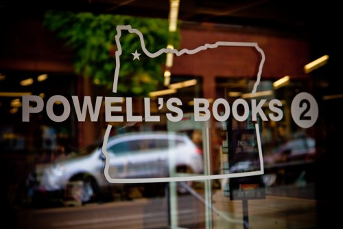 Sign for Powell's Books featuring a map outline of Oregon, reflecting a bookstore window with a city view.
