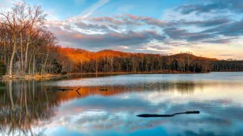 A serene lake at sunset, reflecting colorful clouds and trees along the shore, with a calm water surface.