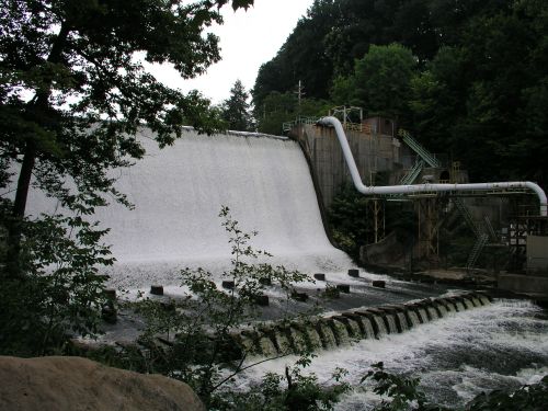 A large waterfall cascades over a dam, surrounded by lush greenery and industrial structures.
