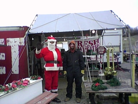 A man in a Santa costume stands next to another man in work attire outside a festive market stall.