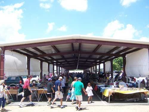 A bustling outdoor market under a large pavilion, with people browsing various stalls and goods on display.