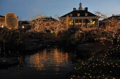 A serene garden illuminated with twinkling lights, reflecting in a pond at dusk, with a charming building in the background.