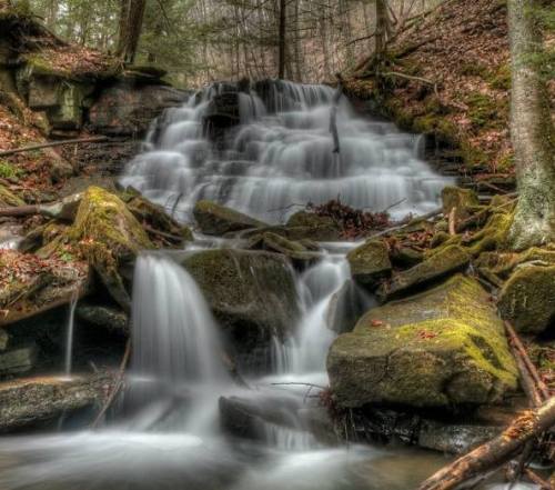 A serene waterfall cascades over moss-covered rocks in a lush, wooded area.