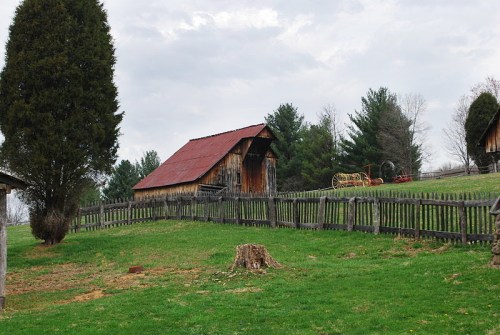 A rustic barn with a red roof sits on a grassy hillside, surrounded by trees and a wooden fence under a cloudy sky.