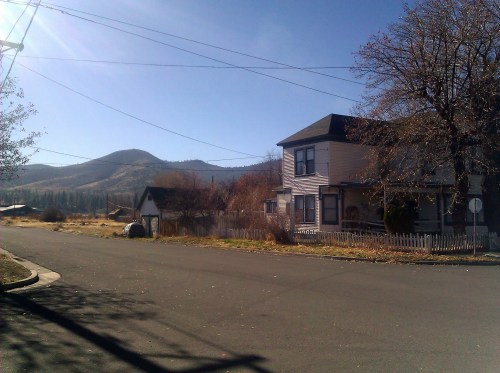 A quiet street scene featuring a house, a small shed, and mountains in the background under a clear blue sky.