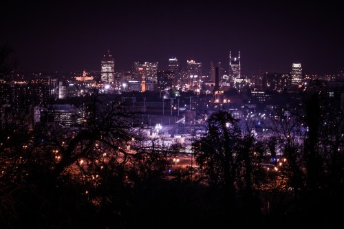 City skyline at night, illuminated buildings and lights, with trees in the foreground creating a silhouette.