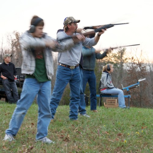 A group of people shooting rifles at a range, wearing ear protection, with trees in the background.