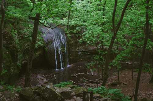 A serene waterfall cascades into a forested area, surrounded by lush green trees and rocky terrain.