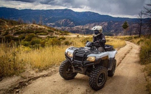 A person rides an ATV on a dirt path surrounded by mountains and autumn foliage under a cloudy sky.