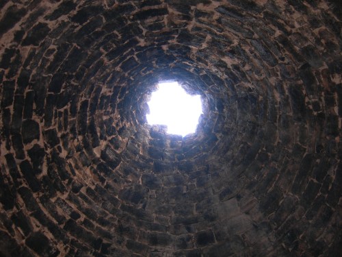 View looking up through a circular stone tower, revealing a bright sky through an opening at the top.