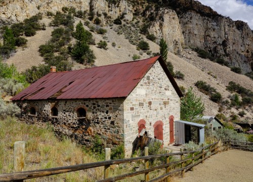 An old stone building with a red roof, surrounded by rocky hills and greenery, set in a rural landscape.