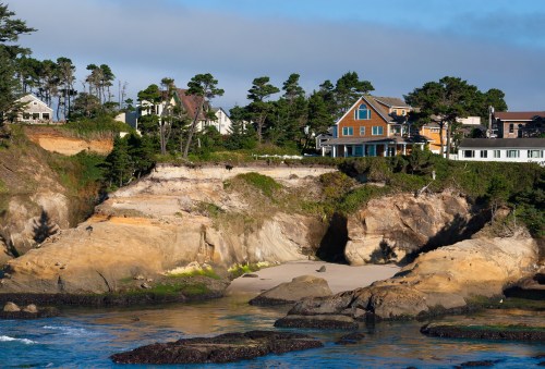 Coastal scene featuring houses on a cliff, sandy beach, and rocky shoreline under a clear blue sky.