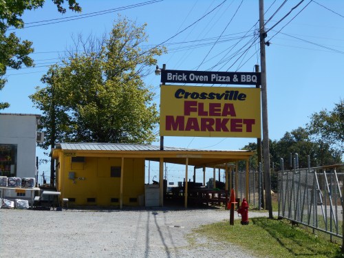 A bright yellow sign for Crossville Flea Market, with a nearby pizza and BBQ restaurant, set against a clear blue sky.