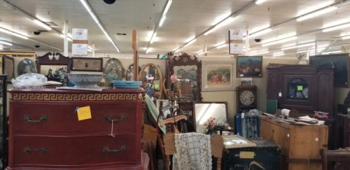 A cluttered antique store filled with various furniture, mirrors, and artwork, with price tags visible.