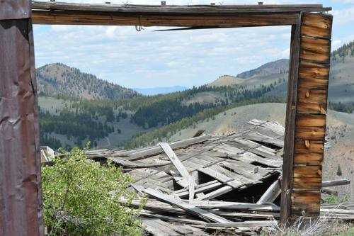 A weathered wooden frame overlooks a collapsed structure and rolling green hills under a cloudy sky.