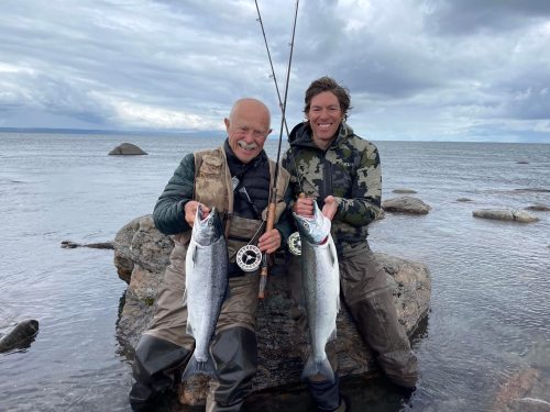 Two men stand on a rock by the water, holding large fish and fishing rods, with a cloudy sky in the background.