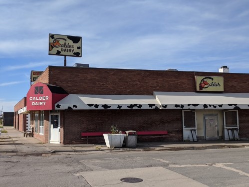 Exterior of Calder Dairy building with a sign, featuring a red awning and a cow-themed design, on a clear day.