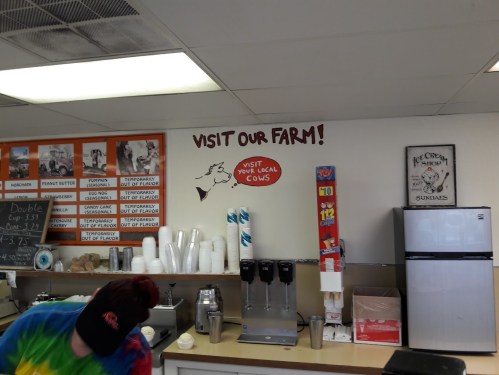 A colorful ice cream shop interior with a "Visit Our Farm!" sign and a worker behind the counter.