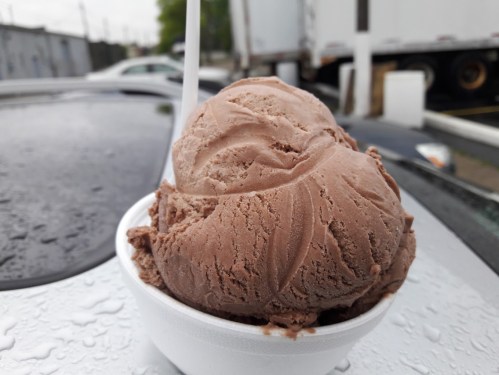A close-up of a cup filled with chocolate ice cream, sitting on a wet surface with a blurred background.