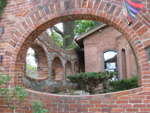 A brick archway frames a garden area with stone accents and a building in the background, surrounded by greenery.