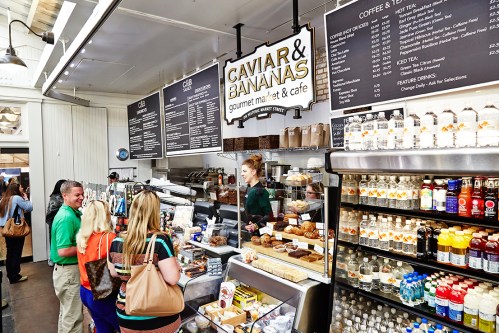 A busy café with a menu board, customers ordering, and shelves stocked with drinks and gourmet items.
