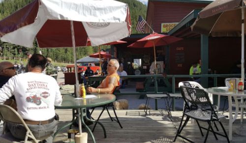 Outdoor café scene with patrons sitting under umbrellas, surrounded by trees and a rustic building in the background.