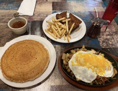 A plate with a large pancake, fries, toast, and a sizzling dish topped with eggs and cheese, with syrup on the side.