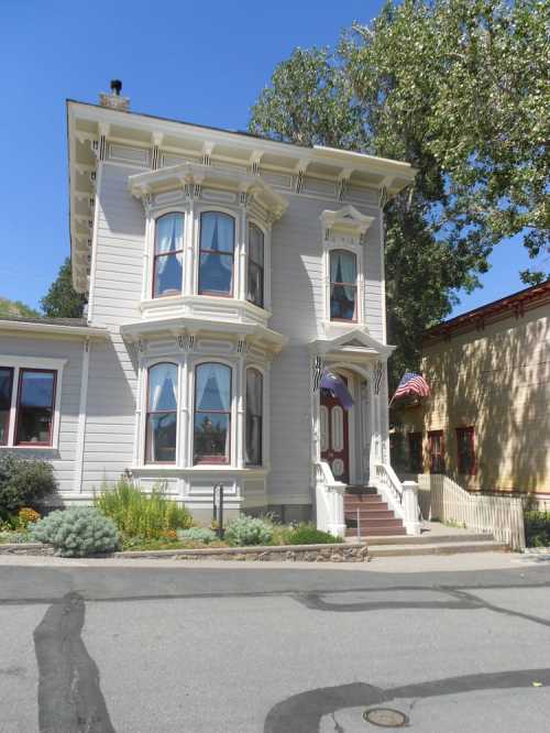 Victorian-style house with bay windows, white exterior, and American flags, surrounded by greenery on a sunny day.