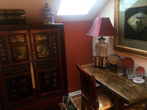 A cozy room featuring a decorative cabinet, a lamp, a small table, and various glassware against a warm-colored wall.