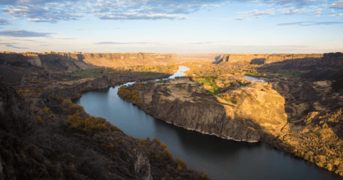 A scenic view of a winding river surrounded by cliffs and lush greenery under a partly cloudy sky.