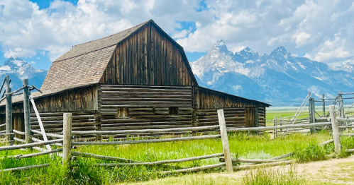 A rustic wooden barn surrounded by green grass, with majestic mountains in the background under a partly cloudy sky.