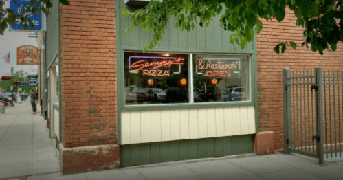 A corner restaurant with a neon sign reading "Sammy's Pizza & Restaurant Open" in a brick building.