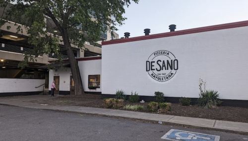 Exterior of Pizzeria DeSano, featuring a white wall with a circular logo, trees, and a nearby parking structure.