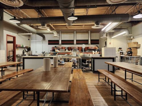 A spacious restaurant interior with wooden tables, industrial decor, and staff working in an open kitchen.