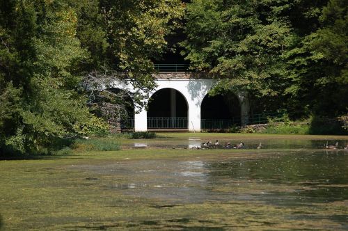 A serene lake scene with a white archway and lush greenery, featuring ducks resting on the shore.
