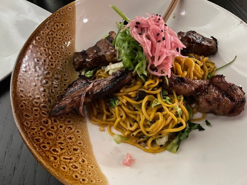 A bowl of grilled meat skewers on a bed of noodles, topped with greens and pickled ginger.