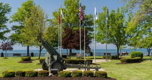 A park with a cannon, flags, and trees, overlooking a lake on a clear day. Benches are visible in the background.