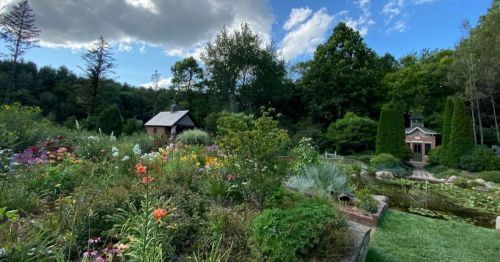 A lush garden filled with colorful flowers, a small house, and trees under a partly cloudy sky.