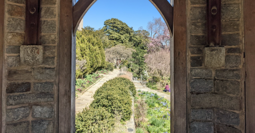 View through an arched stone window, showcasing a lush garden path lined with trees and blooming flowers.
