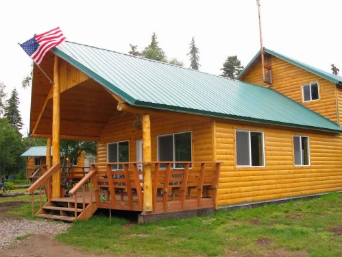 A wooden cabin with a green metal roof, featuring a porch and an American flag, surrounded by trees.