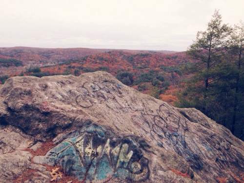 A rocky outcrop with graffiti overlooks a colorful autumn landscape of trees and hills under a cloudy sky.