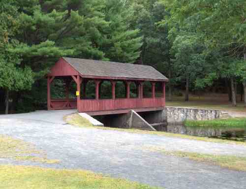 A red covered bridge spans a small pond, surrounded by green trees and a gravel path.