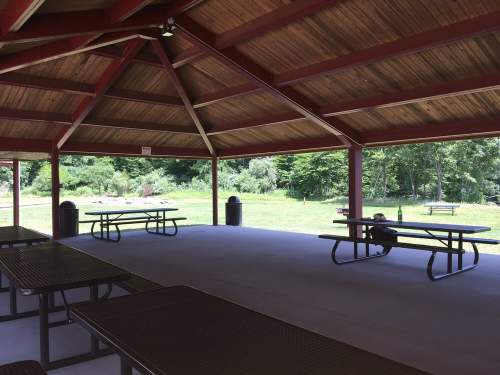 A spacious, open pavilion with wooden beams, empty picnic tables, and a view of greenery outside.