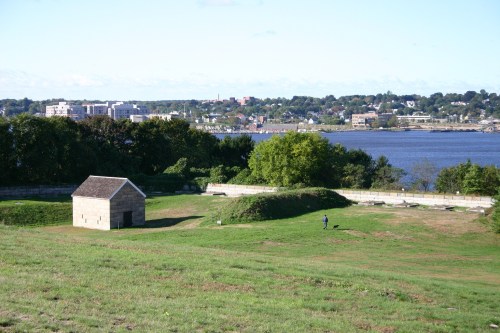 A grassy landscape with a small stone building, overlooking a river and distant city skyline under a clear blue sky.