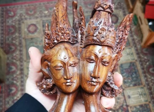 A hand holding a carved wooden sculpture of two faces with ornate headdresses, symbolizing duality or unity.