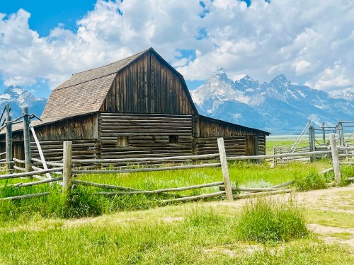 A rustic wooden barn surrounded by a fence, with mountains and a blue sky in the background.